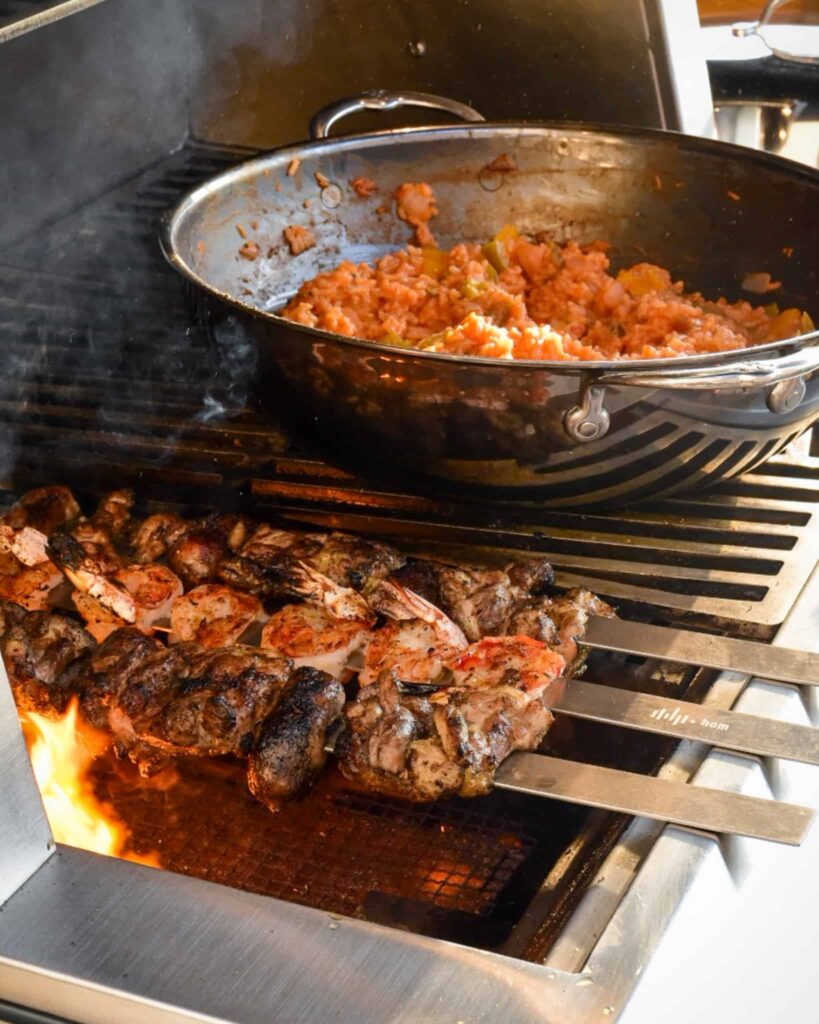 Three skewers of chicken, shrimp and sausage on the grill and a big wok of jambalaya rice and veggies beside it.