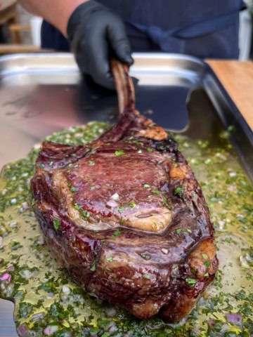 A grilled bison frenched ribeye steak on a tray with a fresh herb sauce.