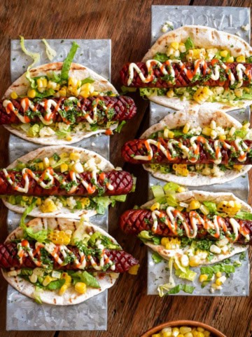 Six crosshatched beef smokies in taco holders dressed with Elote-style corn ingredients.