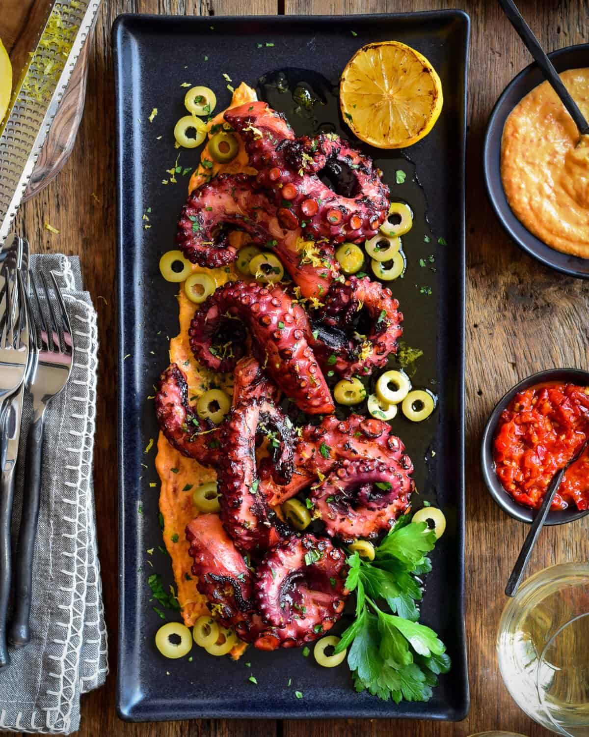 A black rectangular plate with grilled and very tender sous-vide octopus tentacles on a bed of spicy red pepper hummus.