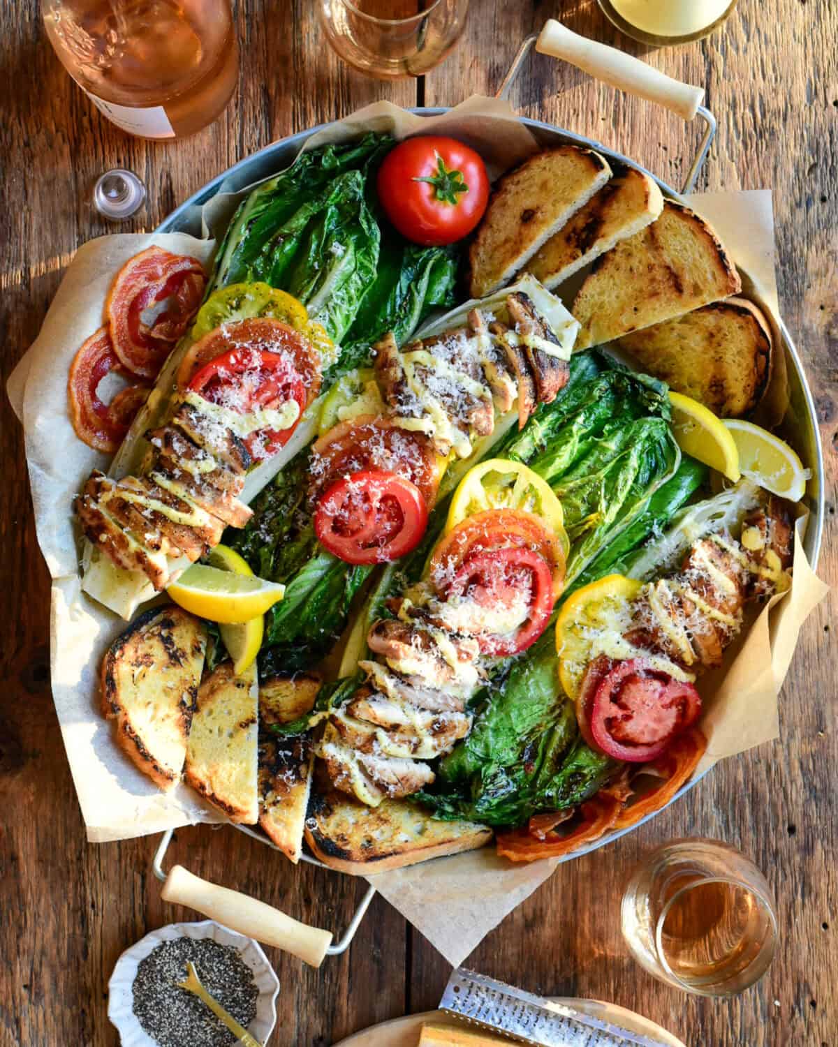 Four grilled romaine halves are topped with tomatoes, pancetta and chicken thighs and drizzled with a Caesar dressing, Parmesan and sourdough toasts in a round serving tray.