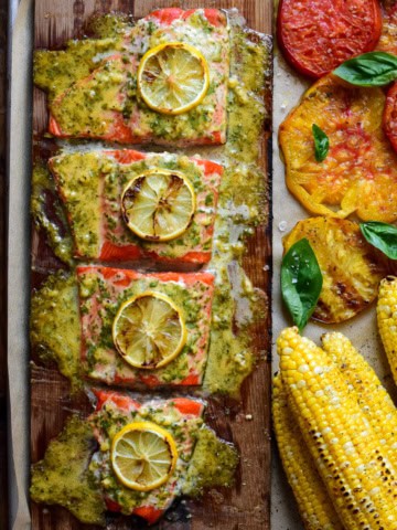 Cedar Plank Sockeye Salmon portioned in four with sauce and lemon slices. Served with grilled corn and tomatoes.