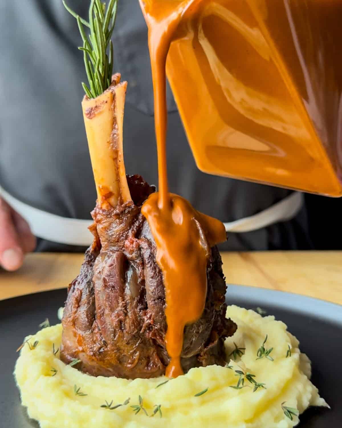 A braised lamb shank placed on a mound of mashed potatoes with sauce poured over it.