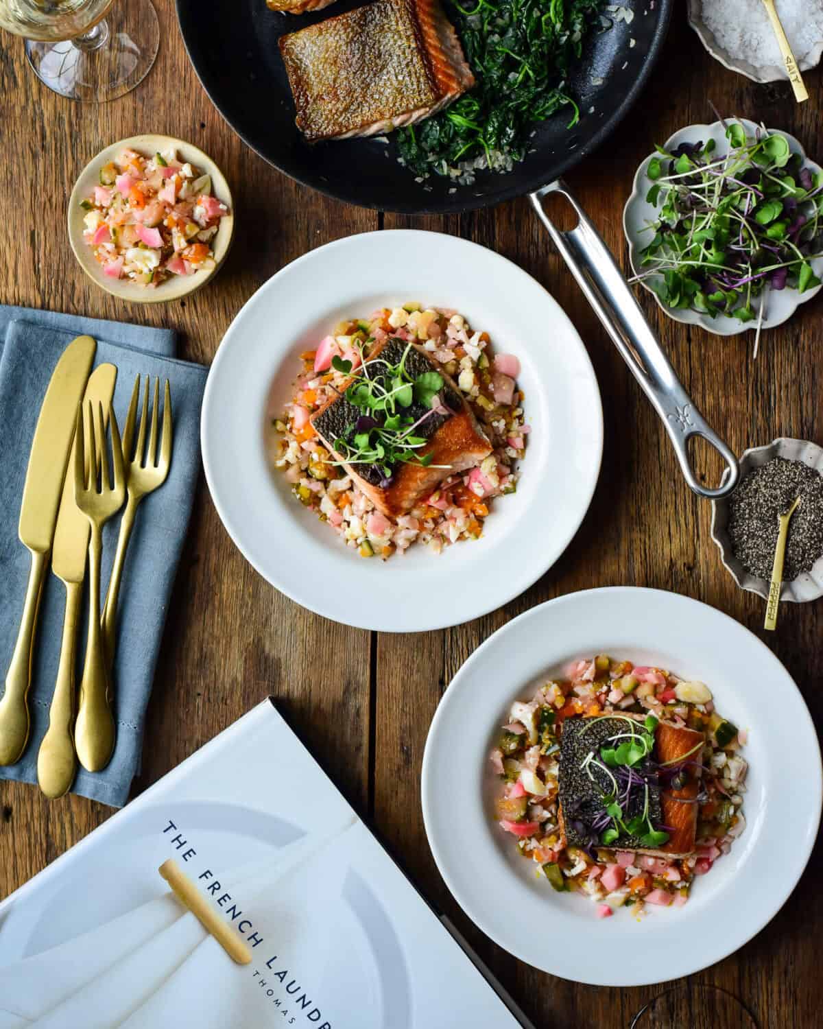 Two plated dishes of Ora King Salmon with Spinach & Chow-Chow. A Thomas Keller skillet and The French Laundry cook book are also on the table.