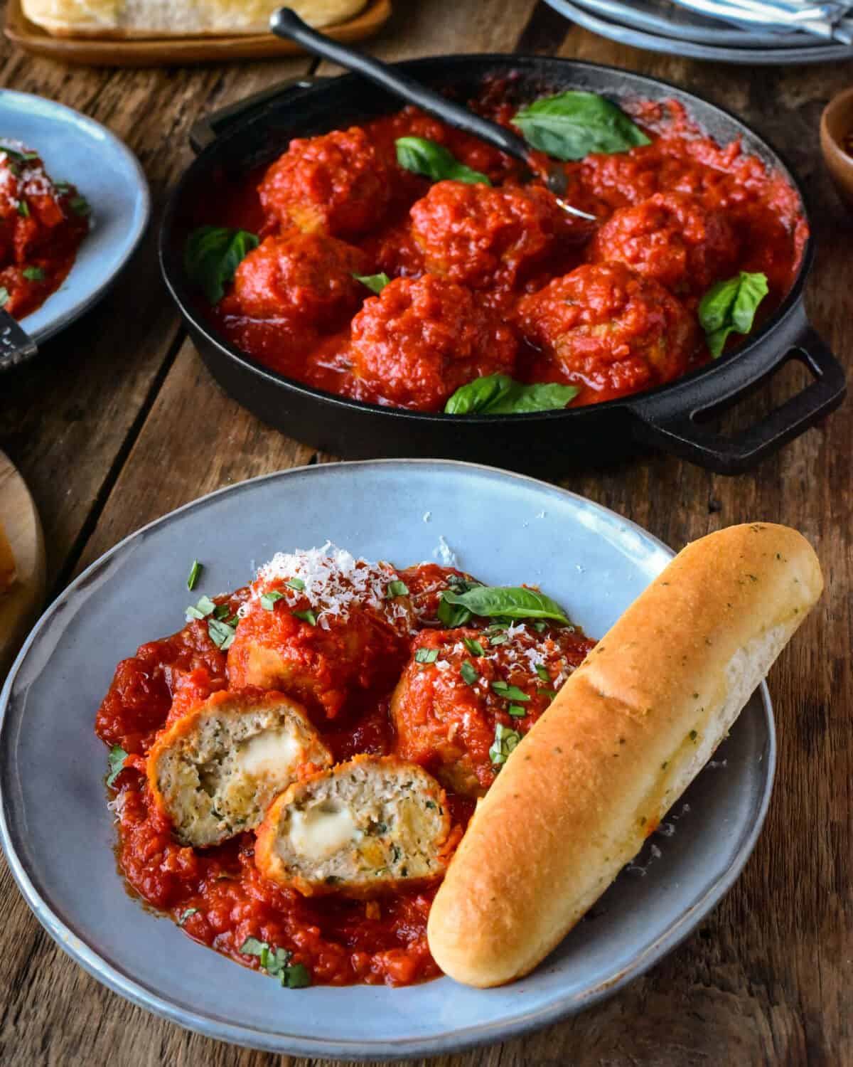 A bowl of Air Fryer Chicken Parmesan Meatballs with a breadstick and a skillet behind with more meatball in sauce.