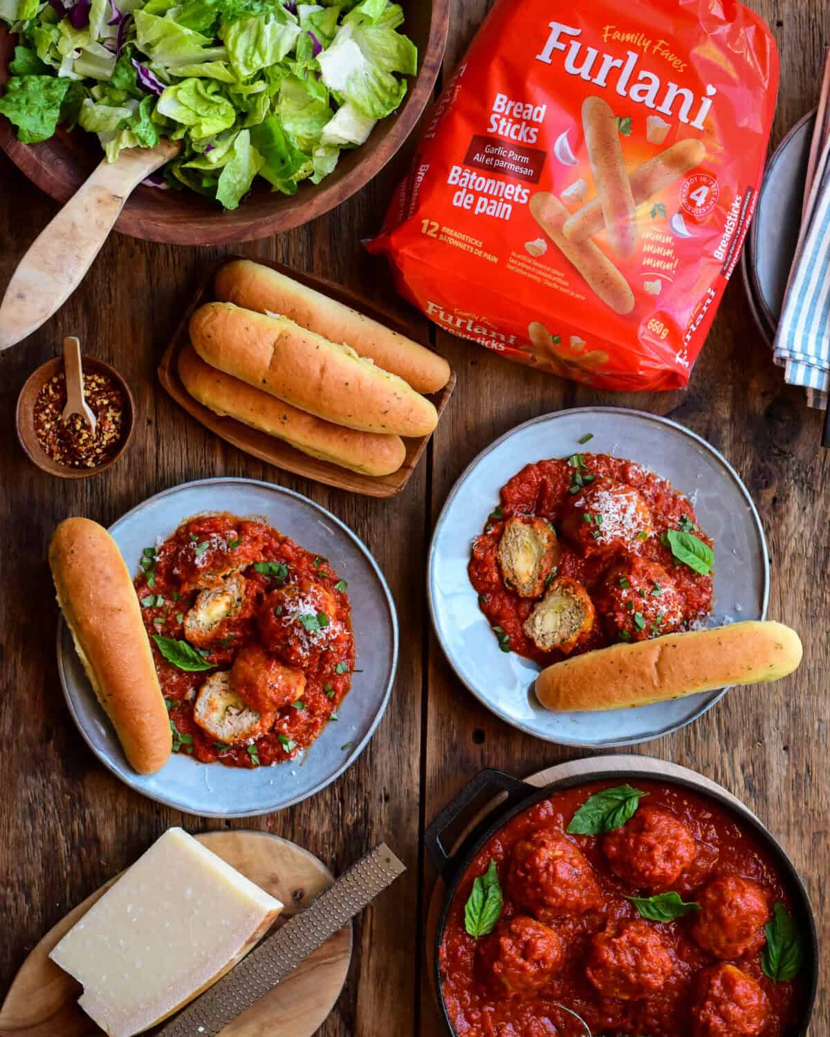 Two plates with meatballs in sauce with breadsticks. The meatballs are cut open and you can see the cheese in the center. Served with a salad grated parmesan and more meatballs in sauce.