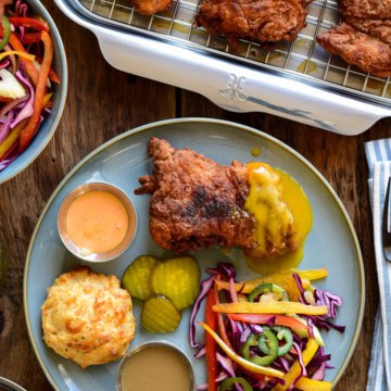A plate with a piece of spicy buttermilk fried chicken served with a psychedelic slaw, cheddar biscuits and three dips.