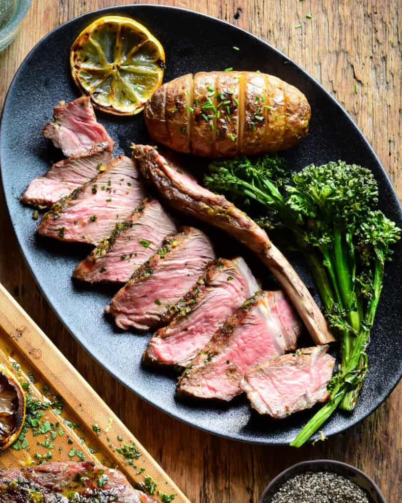 A sliced Tuscan veal chop served on a black plate with roasted broccolini, a roasted potato and a grilled lemon.