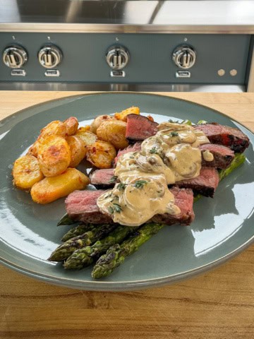 A plate with grilled steak slices overtop asparagus with a mushroom sauce and bomba potatoes.