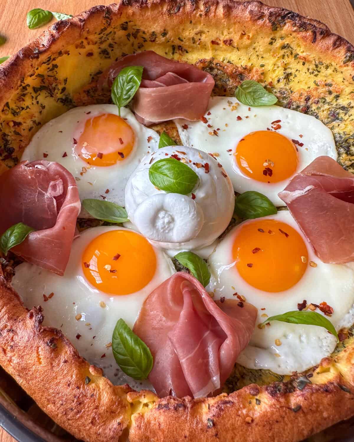 A close up of a savory Dutch baby topped with fried eggs, prosciutto, burrata, basil & chili flakes.