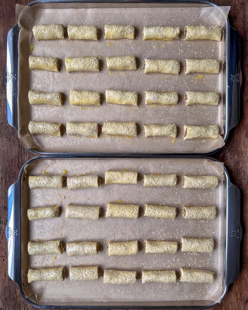 Two sheet pans with rows of sausage rolls brushed with an egg wash and sprinkled with seasame seeds.