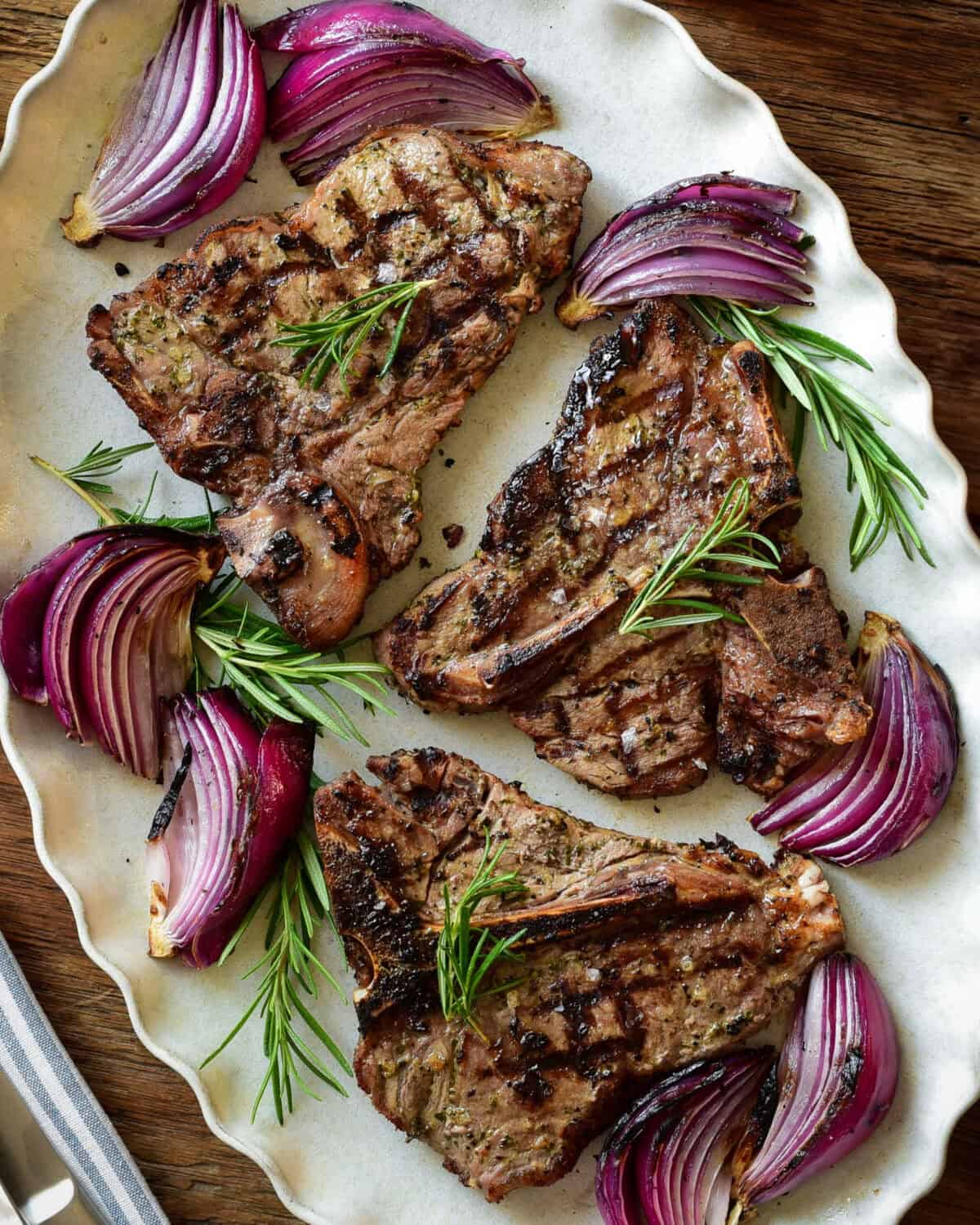 A platter with three grilled veal t-bone chops with grilled red onions and rosemary.