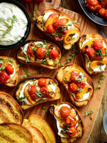 Close up of toasted garlic bread with a layer of whipped feta and topped with 3 grilled peach slices and 3 blistered tomatoes and garnished with fresh basil leaves.