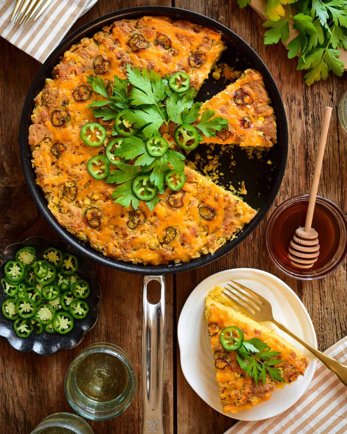 A skillet with a grilled Jalapeño Cheddar Cornbread with a few pieces removed. It's topped with fresh jalapenos and parsley. Served wtih hot honey on the side.