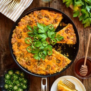 A skillet with a grilled Jalapeño Cheddar Cornbread with a few pieces removed. It's topped with fresh jalapenos and parsley. Served wtih hot honey on the side.