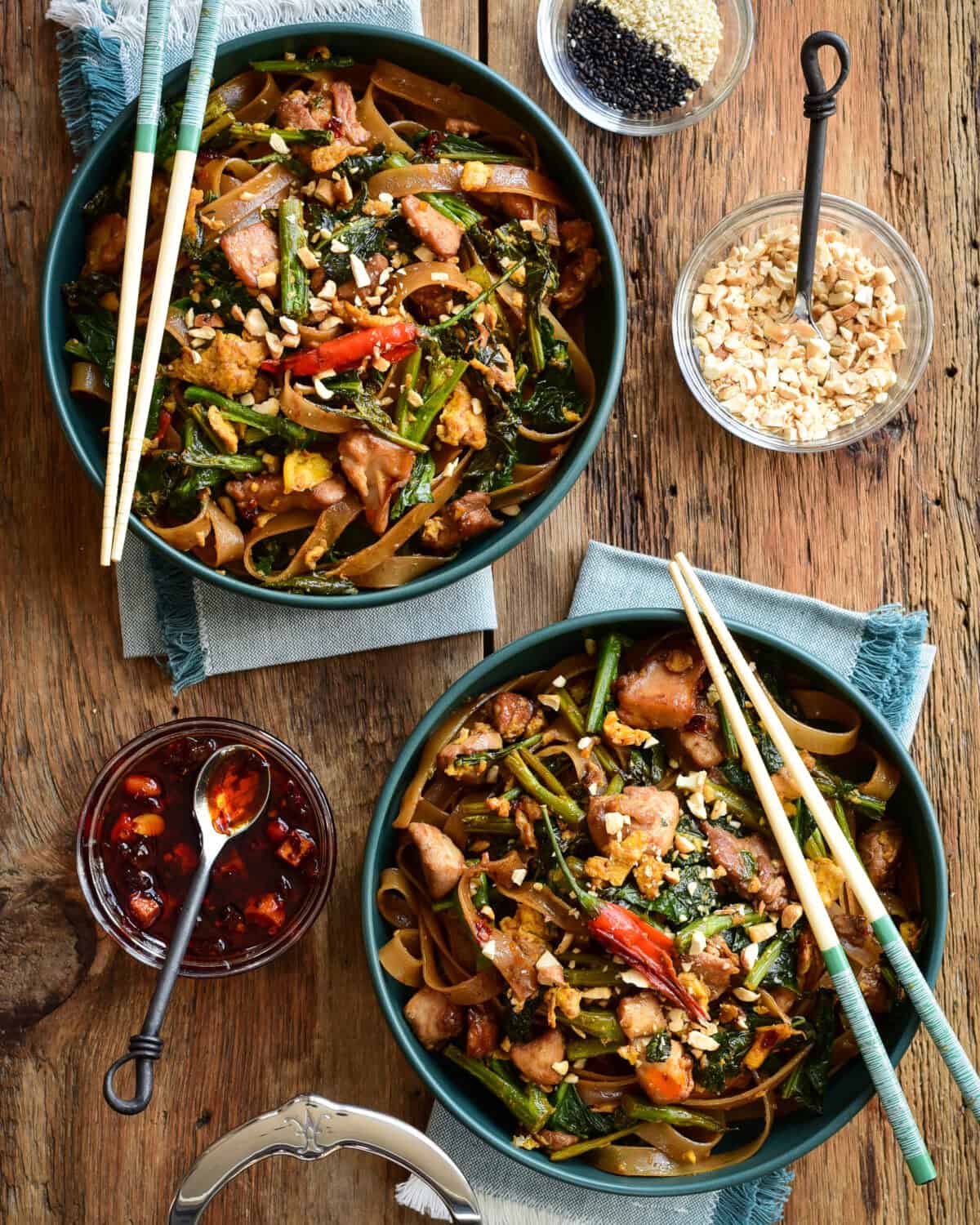 Two bowls of Thai Chicken Stir Fry with Rice Noodles with chopsticks and toppings on the side.
