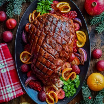 This Festive ham is crosshatched and is plum glazed. It's on platter and is garnished with plums, oranges and festive greenery.