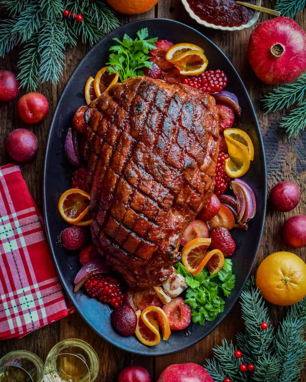 This Festive ham is crosshatched and is plum glazed. It's on platter and is garnished with plums, oranges and festive greenery.
