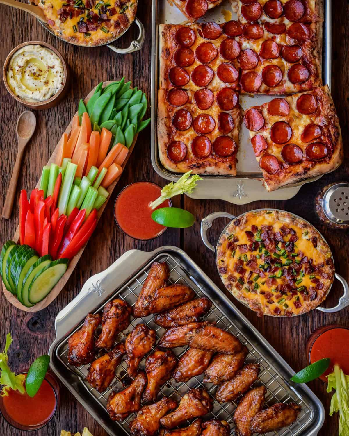 A super bowl spread with an Easy Sheet Pan Pizza with Pepperoni, Crispy and Sticky Chicken, Baked Jalapeno Popper Dip, hummus, fresh veggies and spicy Ceasars.