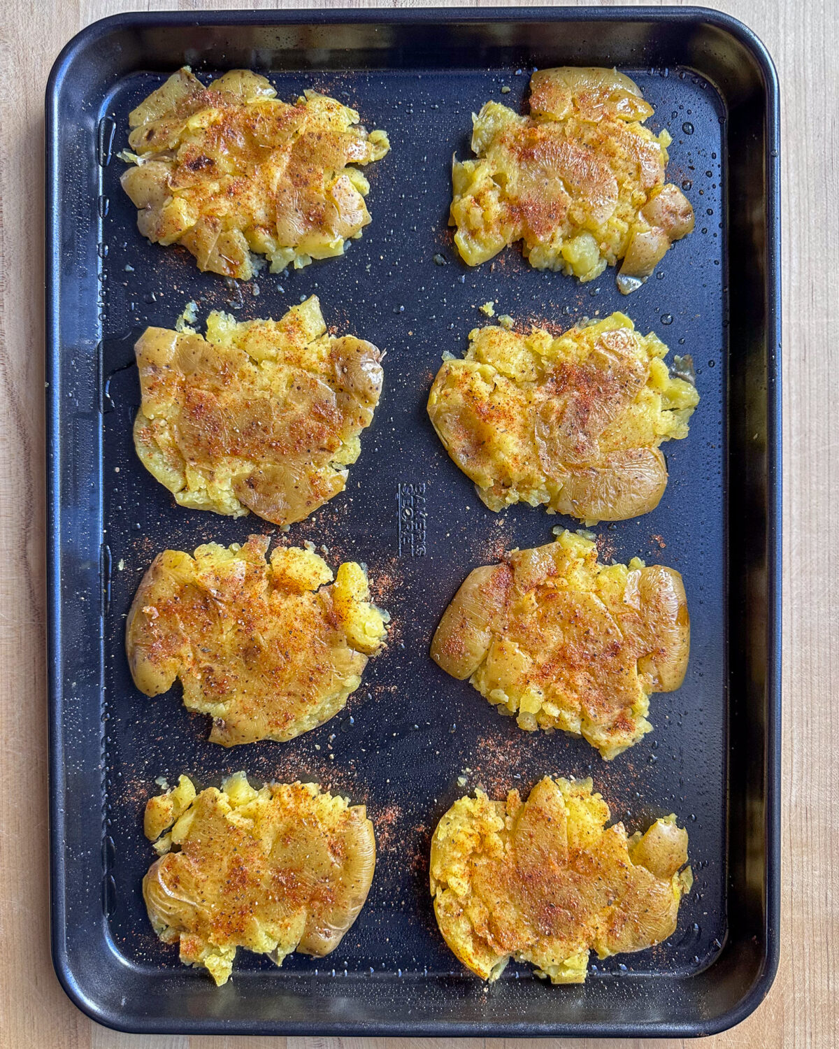 A sheet pan of eight smashed yellow potatoes with seasoning.