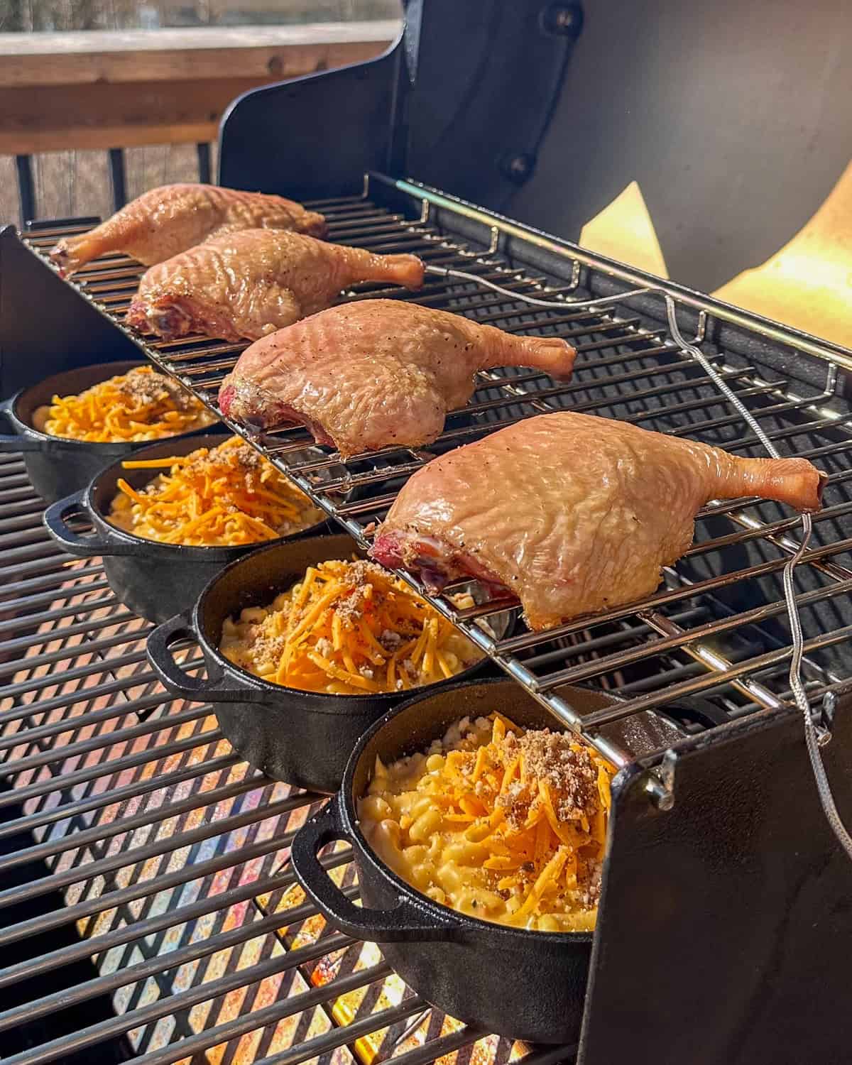 Four smoked duck legs on the top rack of a smoker with four mac and cheese dishes below on the bottom rack.