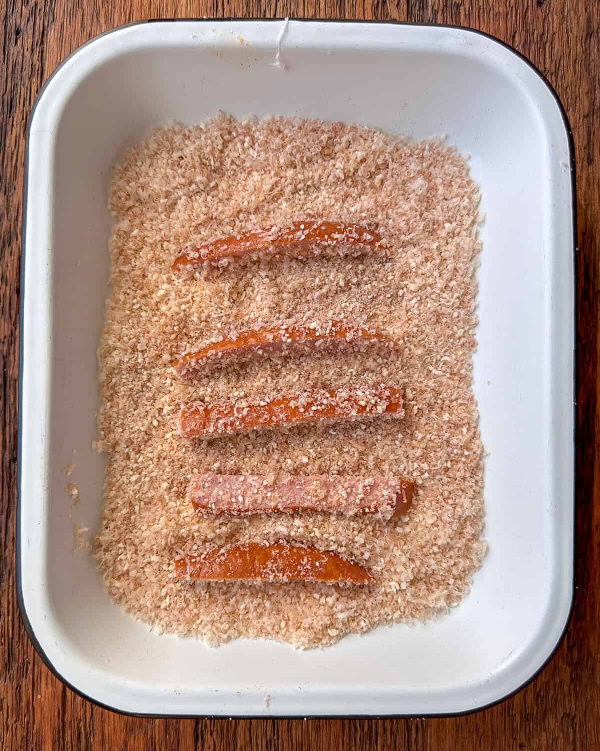 A tray of gluten-free breadcrumbs with five kielbasa fries getting coated in it.