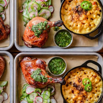 Two trays each with a smoked duck leg, served with a Charred Scallion Chimichurri, mac & cheese, and a cucumber salad.