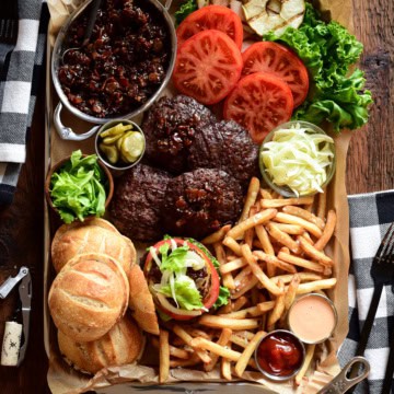 A serving tray with Espresso Bacon Jam Burgers with toppings, fries and a pot of smoked Gouda Sauce.