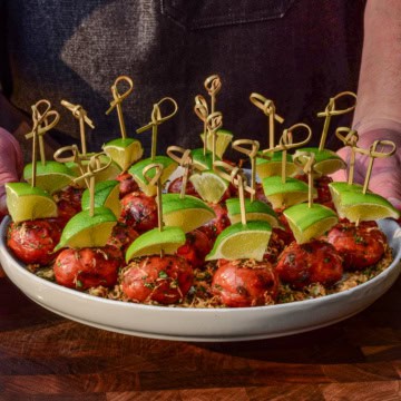 A platter of lil smokies skewered with lime wedges and coated with an elote topping.