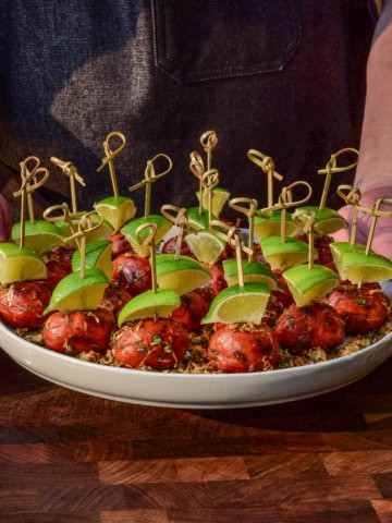 A platter of lil smokies skewered with lime wedges and coated with an elote topping.