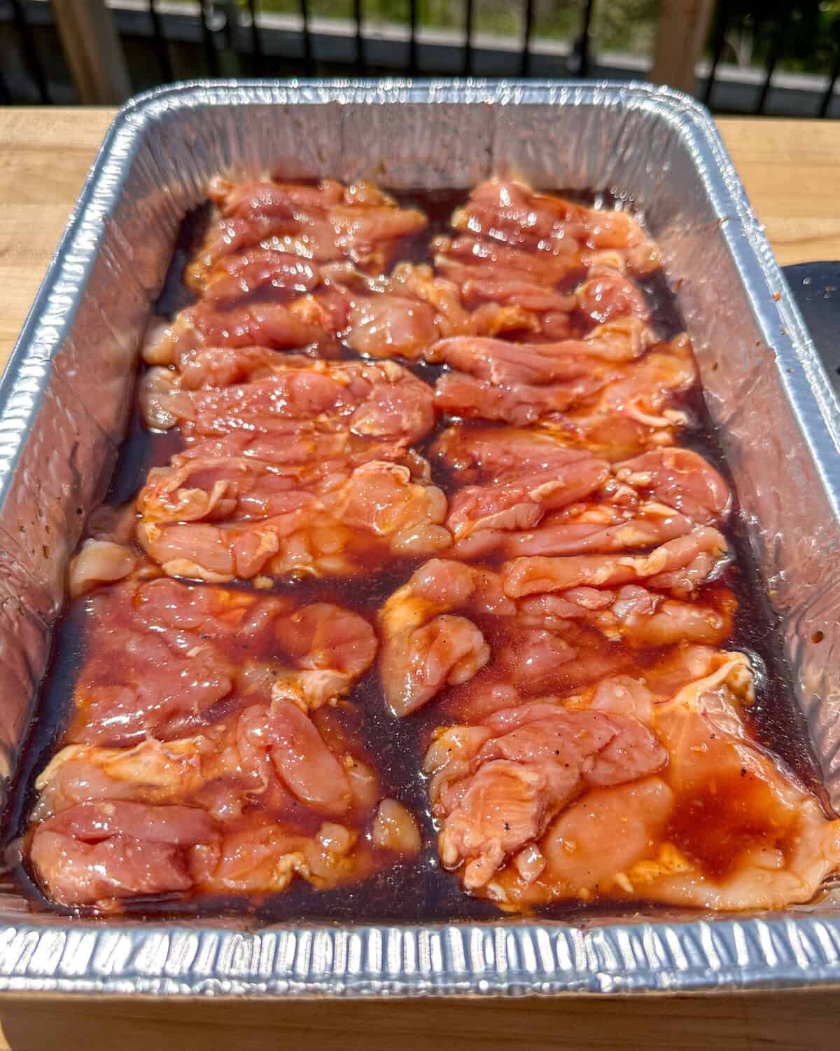 A foil tray with chicken thighs marinating in a teriyaki marinade.