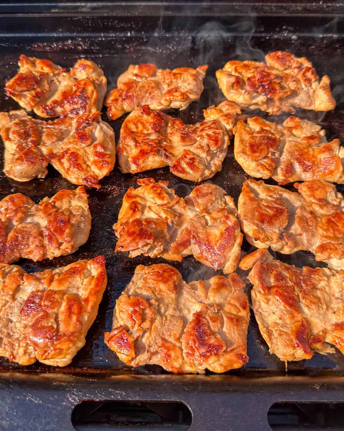 Twelve golden brown teriyaki-marinated chicken thighs on a griddle for chicken sliders.