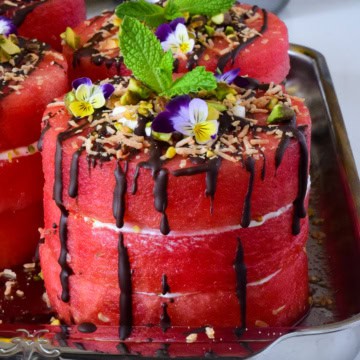A close-up of mini watermelon cakes with coconut whipped cream, dark chocolate drizzle and topped with pistachios, toasted coconut, mint and edible flowers.