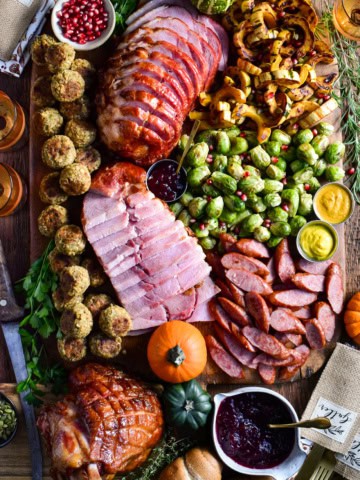 Slices of festive ham and turkey kolbassa are artfully arranged on a board with roasted Brussels sprouts, caramelized delicata squash, stuffing balls, cranberry sauce, and soft dinner rolls. 
