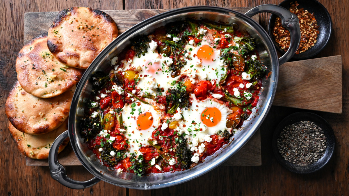 An oval serving dish with a cherry tomato and broccolini shakshuka. Served with mini pita breads and crushed chili peppers.