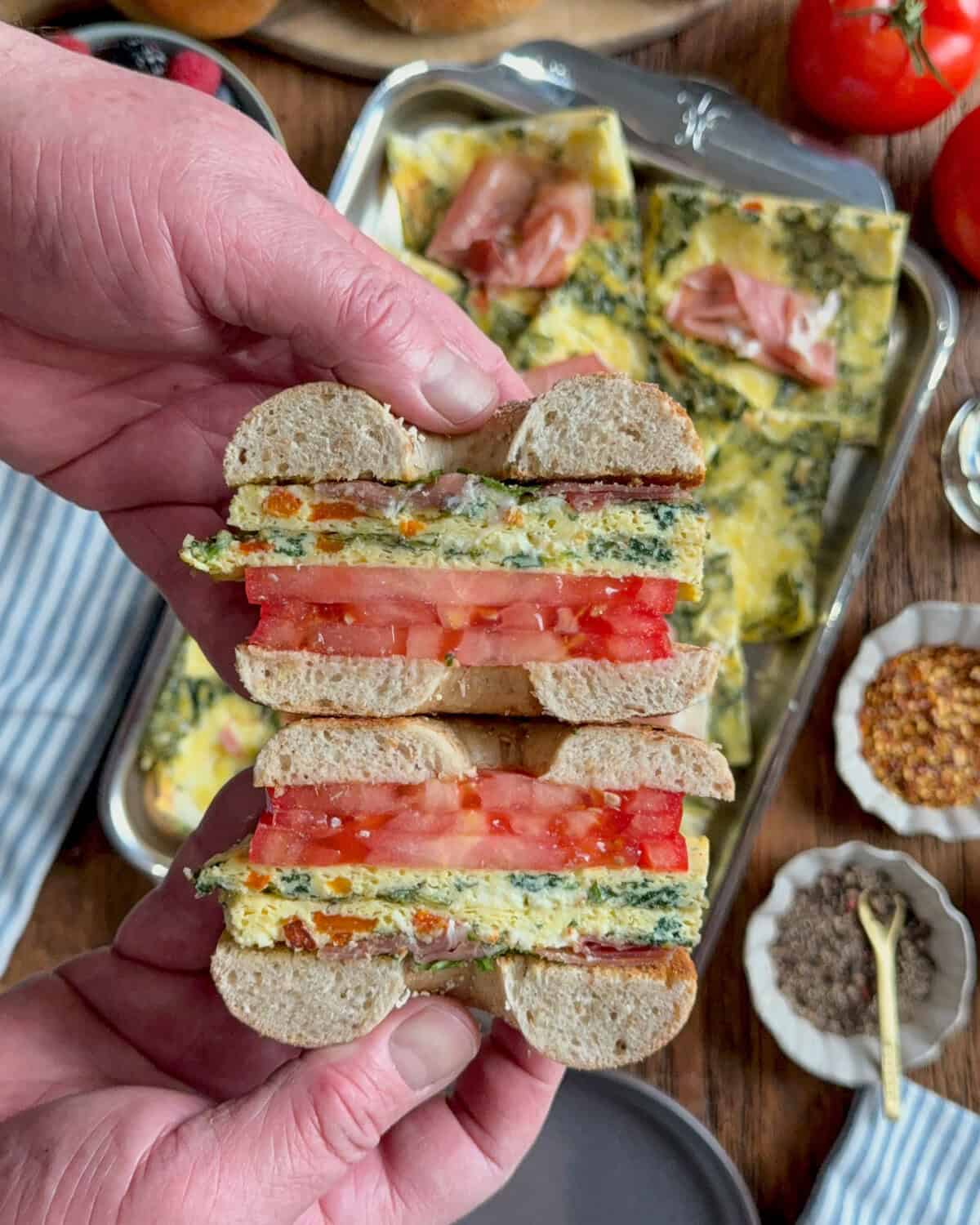 A cross section of a bagel sandwich with sheet pan eggs, tomatoes, prosciutto and arugula.