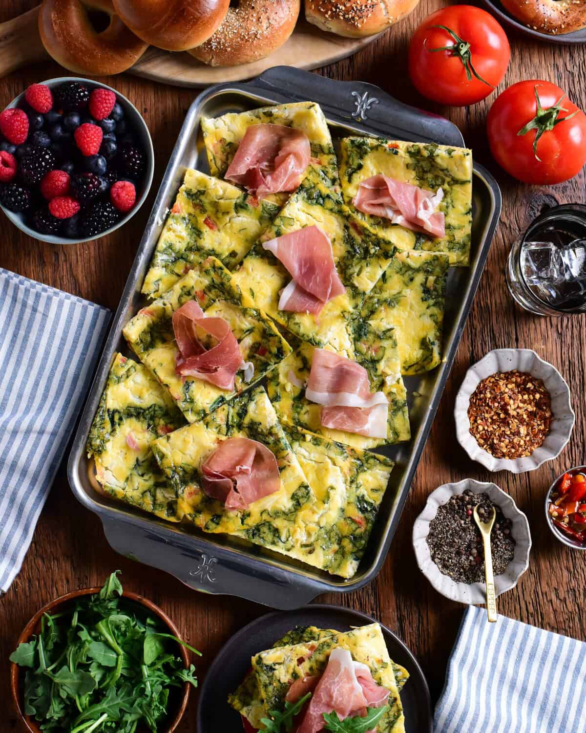 Sheet pan eggs cut into squares, served with prosciutto slices, bagels, arugula, and berries.