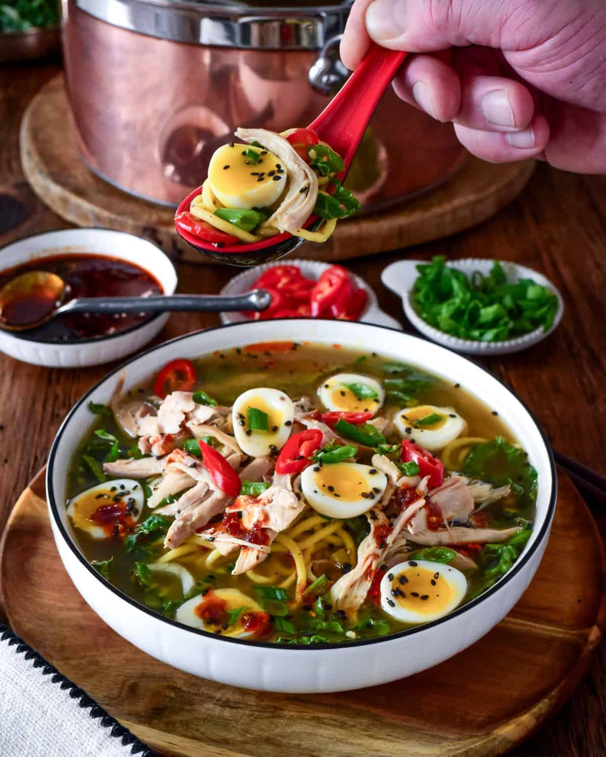 A bowl of ginger chicken ramen soup with bok choy, ramen-style noodles and jammy quail eggs. A spoonful of soup is being lifted up.