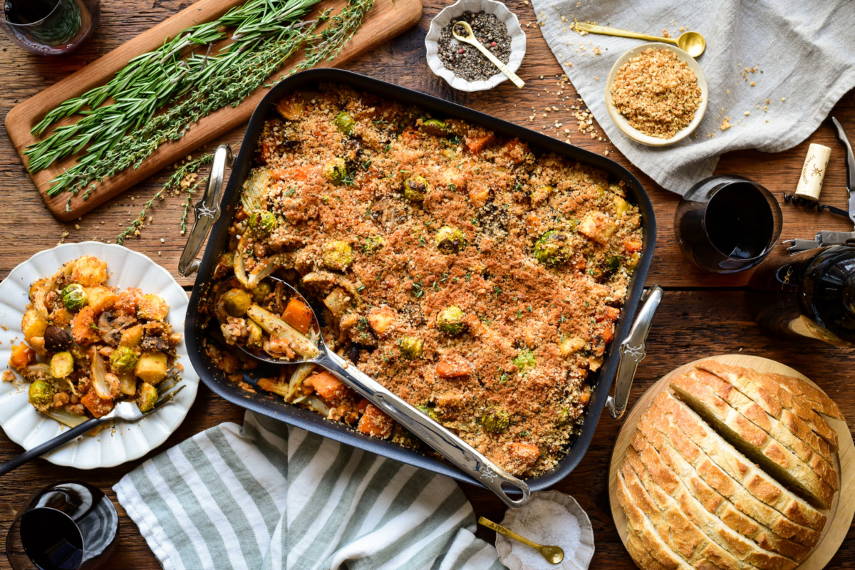 A winter vegetable cassoulet in a roaster with a serving spoon. a portion is served on a plate and is served with extra breadcrumb toppings and sourdough bread.