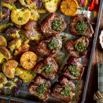 Garlic and anchovy lamb chops with warm salsa verde, served on a stainless-steel griddle with smashed baby potatoes, griddle peppers, and onions.