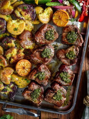 Garlic and anchovy lamb chops with warm salsa verde, served on a stainless-steel griddle with smashed baby potatoes, griddle peppers, and onions.
