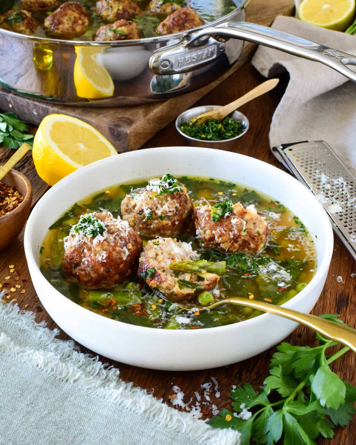 A bowl of chicken meatballs with spring vegetables in a lemon brodo and a pot in the background with more soup.