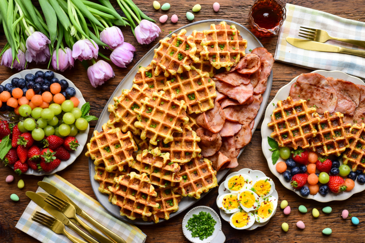 An Easter brunch of cornmeal waffles made with ham and cheddar and served with fruit, extra slices of ham, boiled eggs and syrup.