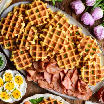 A platter with cornmeal waffles made with ham and cheddar and served with extra old-fashioned ham slices, hard-boiled eggs, fruit, and maple syrup.