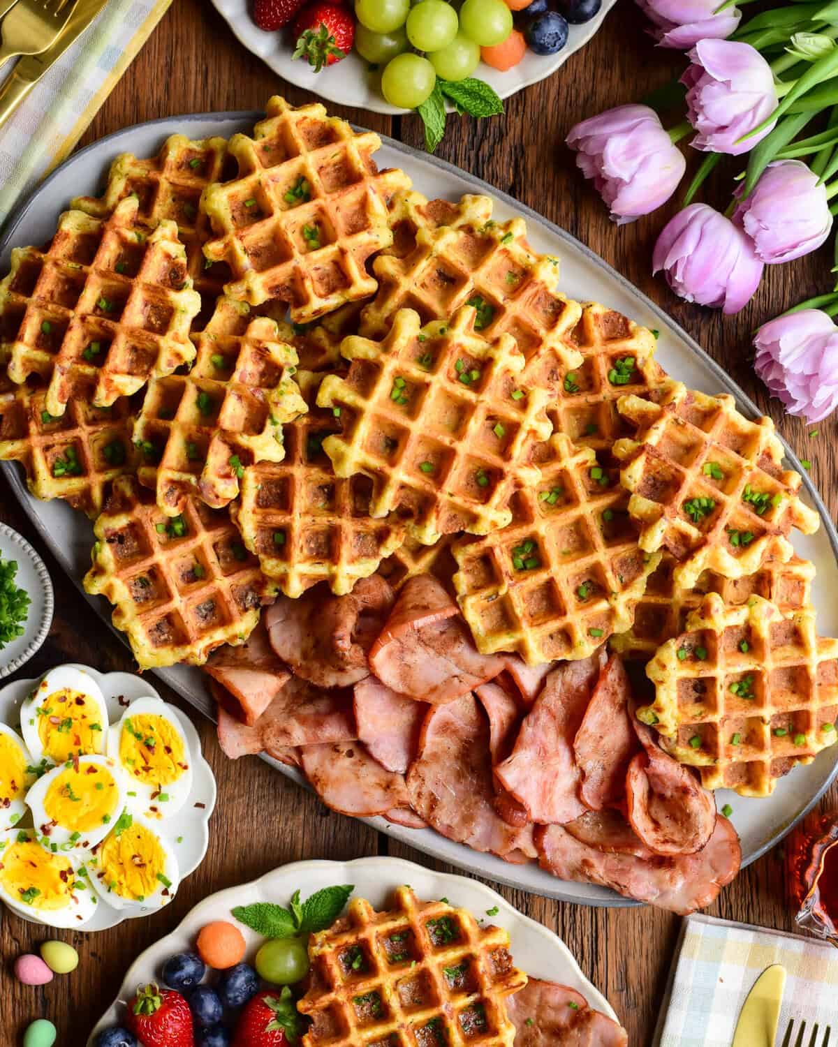 A platter with cornmeal waffles made with ham and cheddar and served with extra old-fashioned ham slices, hard-boiled eggs, fruit, and maple syrup.