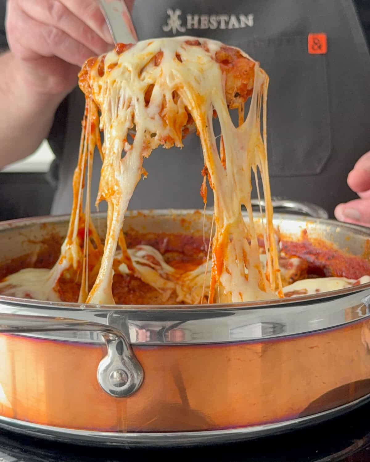 Lifting out a chicken Parmesan thigh from the pan with a cheese pull.