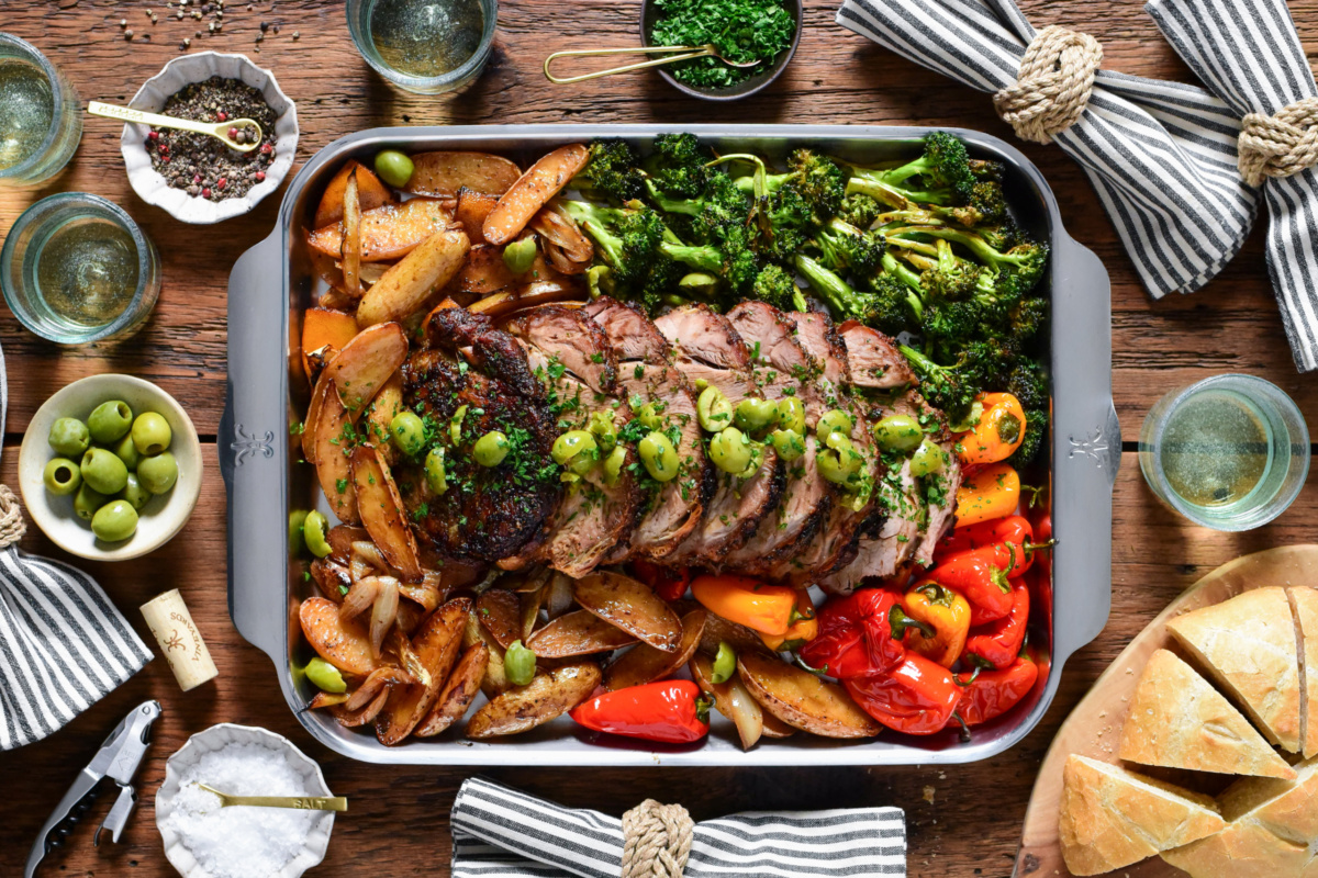 A sheet pan with a sliced oven roasted pork butt served with crushed olives and parsley. Served with roasted fingerling potatoes, peppers, broccoli and bread.