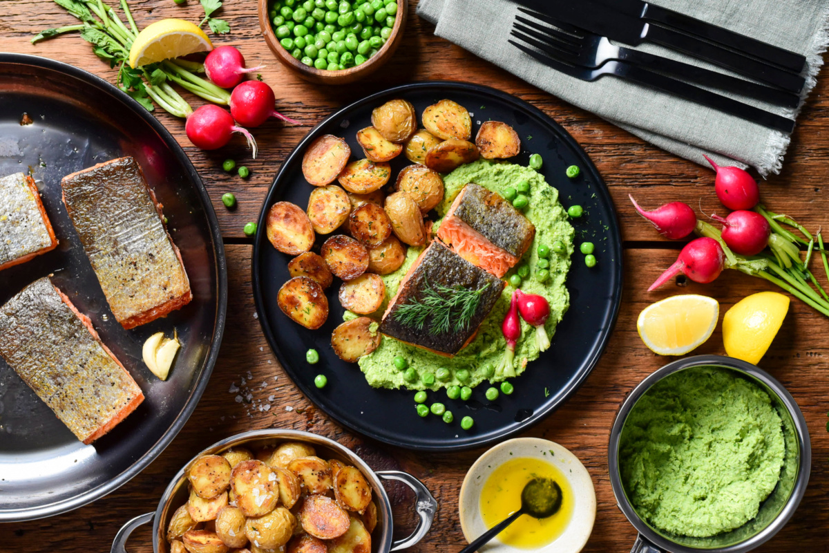 A piece of salmon is cut open and served over a pea puree and potatoes. Extra crispy skin salmon, pea puree, roasted potatoes and lemon oil surround the serving.