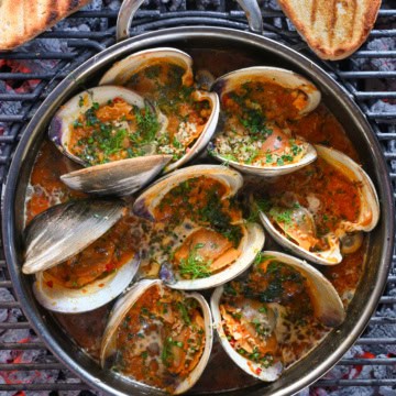 A pot of grilled clams casino in a Calabrian chili sauce over a BBQ with some grilled toast.