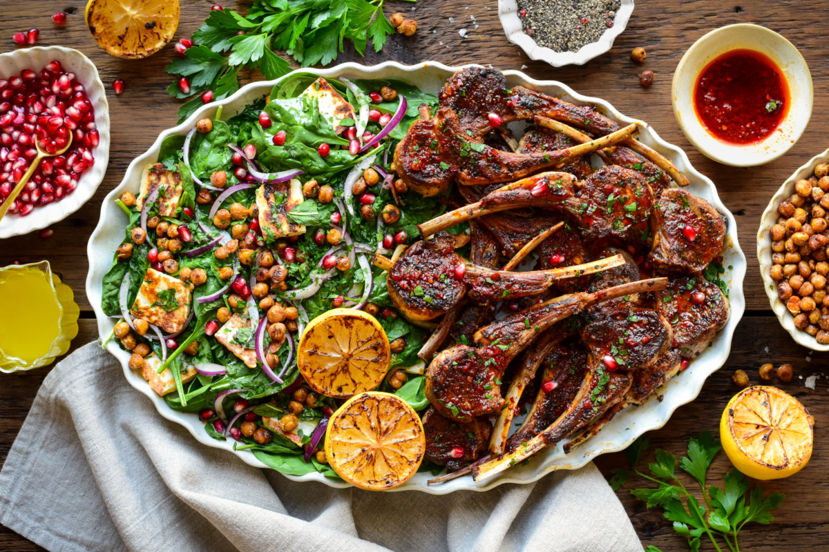 Harissa Lamb Chops with Crispy Chickpea & Halloumi Salad served family style on a platter. Served with extra dressing, pomegranate arils and chickpeas.